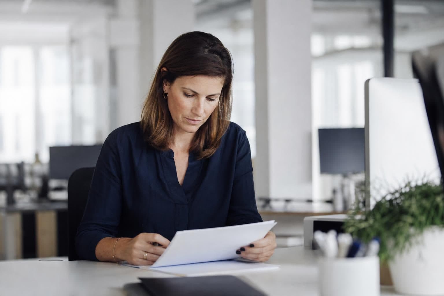 Une femme est assise à un bureau dans un open space lumineux, en train de lire et manipuler des feuilles de papier.